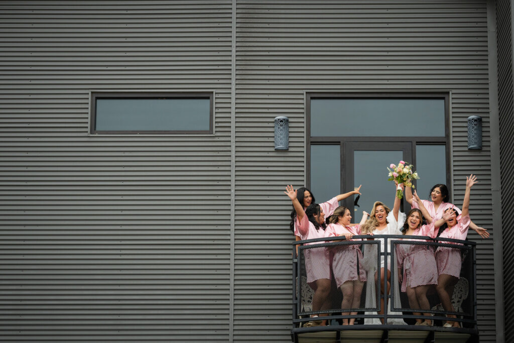 San Antonio wedding photographer captures bride and bridesmaids celebrating in pink robes on balcony during wedding morning getting ready photos